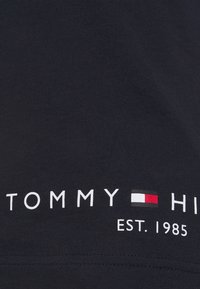 Textil trička s nápisem "TOMMY HILFIGER EST. 1985" a značkovým červenobílým černým obdélníkovým logem mezi slovy.