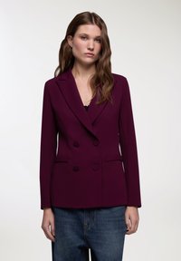 Blazer doppiopetto di un viola scuro con rever a punta, due tasche frontali e una texture liscia, indossato su jeans in denim.