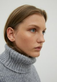 Hellgrauer, gerippter Rollkragenpullover aus weichem, strukturiertem Stoff. Nahaufnahme des Profils eines Modells, das glatte Haut und ein natürliches Make-up zeigt.
