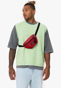 Punainen vyölaukku vetoketjulla, käytetään ristiin yli vaaleanvihreän tekstuurisen oversized paidan ja vaaleansiniset farkut. Musta säädettävä hihna.