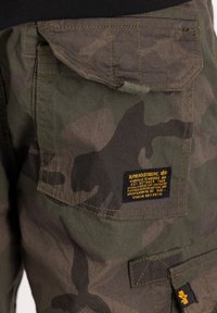 Shorts cargo camouflages dans des tons verts, avec plusieurs poches, des coutures renforcées et une étiquette de marque visible avec du texte jaune.