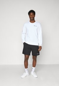 Weißer Langarm-Sweatshirt mit besticktem Logo, kombiniert mit schwarzen Shorts und weißen Sneakers. Enthält weiße Crew-Socken mit Logo.