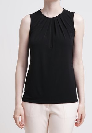 Femme portant un haut noir sans manches avec des plis près de l'encolure et un pantalon de couleur claire sur un fond uni.