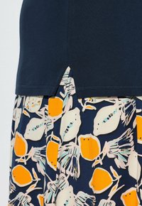 Marineblaues Hemd mit kleinem Seitenschlitz, getragen über einem Rock mit einem abstrakten Muster aus orangefarbener und weißer Frucht auf dunkelblauem Hintergrund.