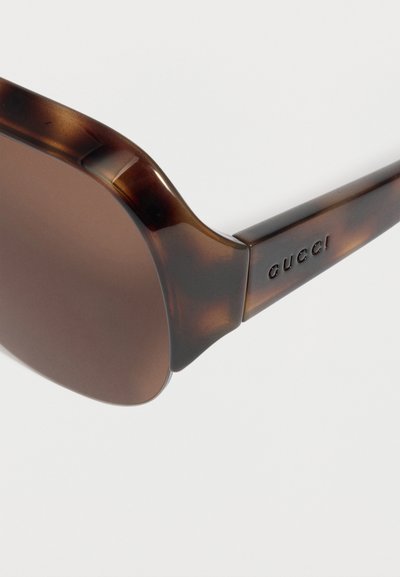 Γυαλιά ηλίου με καφέ πλαίσιο χελώνας, καφέ φακούς και τη χαραγμένη λέξη «Gucci» στο μπράτσο, ορατά σε λευκό φόντο.