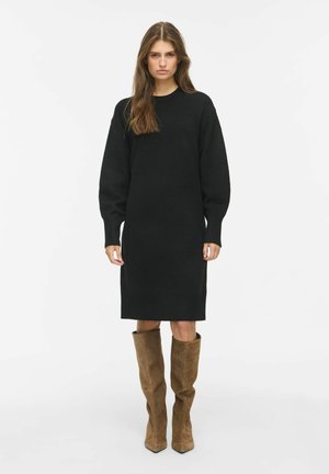 Young woman standing wearing a black long-sleeve knit dress and knee-high brown suede boots against a white background.