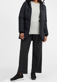 Svart puffjacka med huva och dragkedja, kombinerad med en ljusgrå tröja och svarta byxor med vidben, kompletterad med svarta sneakers.