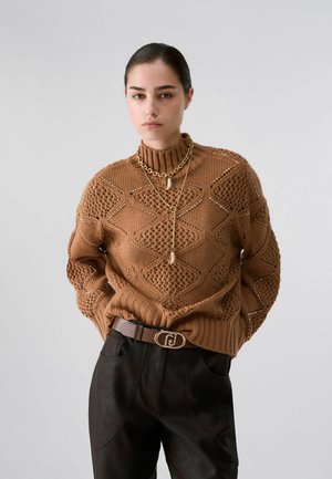 Jeune femme aux cheveux foncés portant un pull en maille marron à motifs, des colliers dorés en couches, un pantalon noir et une ceinture marron avec une boucle dorée.