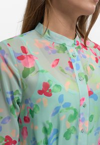 Tunn, ljusblå blus med ett blommönster i röd, rosa, grön och blå. Har en mandarinkrage och knappar fram.