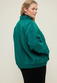 Teal fleecová bunda s vysokým límcem, spadlými rameny a žebrovaným lemem. Modelka má na sobě černou žebrovanou sukni. Jednoduchý, oversize design.
