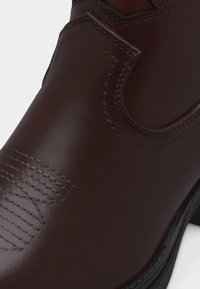 Κοντινό πλάνο σκούρου καφέ δερμάτινου μποτακιού με διακοσμητική ραφή και θηλιά στο πλάι, που δείχνει λείο υλικό και μαύρη άκρη σόλας.