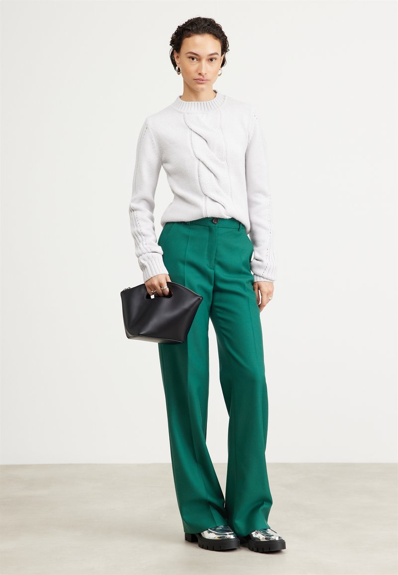 Maglione grigio chiaro lavorato a maglia con dettagli a coste, abbinato a pantaloni verdi a vita alta. La modella tiene una piccola borsa nera. Scarpe con tacco a pianta argentata.