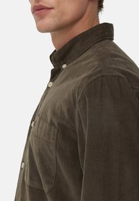 Brązowa koszula z korduroju z kołnierzykiem zapinanym na guziki i przednią kieszenią. Charakteryzuje się teksturowanym materiałem i jasnymi guzikami, przedstawiona z boku.