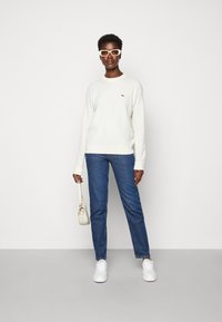 Cream sweater with ribbed cuffs, light denim jeans, white sneakers, and a small cream handbag; features logo on the sweater.