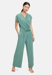 Combinaison drapée vert menthe avec des manches courtes, des jambes larges, des plis à l'avant et une coupe décontractée. Portée avec des talons ouverts de couleur claire.