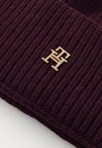 Bonnet en maille côtelée bordeaux avec un accent logo en métal doré. Le tissu semble doux et texturé, mettant en valeur un design ajusté.