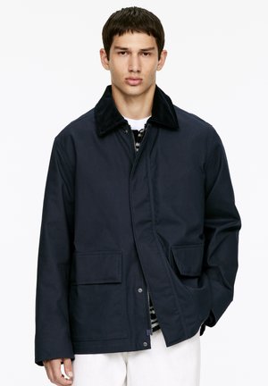 Marineblaue Jacke mit einem Cordkragen, zwei vorderen Taschen und einem Reißverschluss. Das Material wirkt glatt und hat einen lässigen Schnitt.