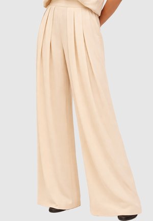 Beige hoch taillierte, weite Hose mit vorderen Falten, getragen mit schwarzen Schuhen und einem passenden beigen Oberteil vor grauem Hintergrund.