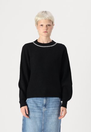 Black ribbed sweater with a rounded neckline featuring a pearl-like accent. Long sleeves and relaxed fit, styled with a denim skirt.