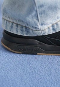 Melnas kedas ar teksturētu auduma augšdaļu un gumijas zoli. Redzamais Adidas logotips sānu daļā. Kombinē ar džinsiem ar gaiši zilu denimam līdzīgu apdari.