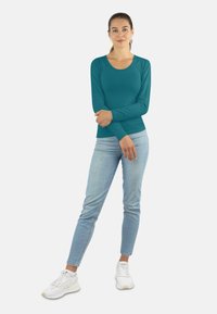 Langärmliges, türkisfarbenes Shirt mit Rundhalsausschnitt, kombiniert mit hellblauen Slim-Jeans und weißen Sneakers, Stehpose vor einem einfarbigen Hintergrund.