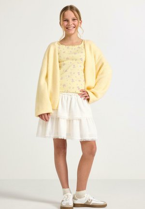 Smilende pige med lyst hår i hestehale iført gul blomsterbluse, gul cardigan, hvid lagdelt nederdel, hvide sokker og beige sneakers.