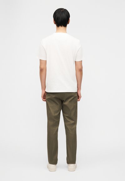 Άνδρας όρθιος με την πλάτη προς τα πίσω, φορώντας ένα απλό λευκό μπλουζάκι, ελιά πράσινο παντελόνι και εκρού αθλητικά παπούτσια σε απλό φόντο.