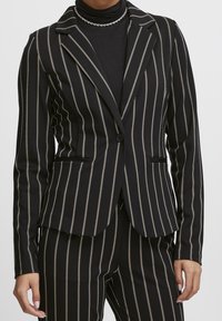 Blazer à rayures noires et crème avec une coupe ajustée, des revers crantés, une fermeture à un bouton et des poches latérales. Tissu à texture lisse.