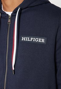 Felpa con cappuccio blu navy con chiusura con zip, dotata di un grande patch in gomma con la scritta "HILFIGER" sul petto e cordini a contrasto.
