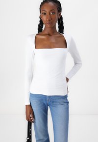 White long-sleeve top with a square neckline, made from a smooth, stretchy fabric. Paired with light blue jeans, features subtle texture.