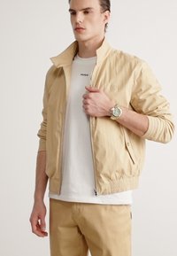 Giacca leggera beige con colletto alto, vita elastica e tasche laterali, indossata sopra una t-shirt bianca. Presenta un'applicazione del logo.