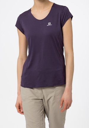 Kobieta w fioletowej koszulce Salomon z krótkim rękawem i beżowych spodniach, stojąca na białym tle.