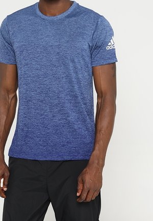 Homme portant un t-shirt Adidas bleu chiné à manches courtes avec le logo sur la manche gauche et un pantalon noir, debout devant un fond blanc.