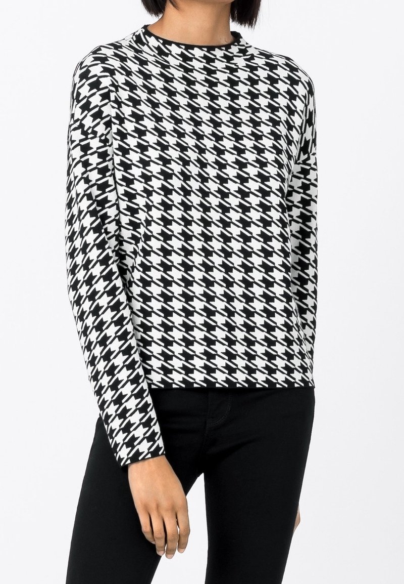 Femme portant un pull à manches longues à motif pied-de-poule noir et blanc et un pantalon noir, debout avec un bras détendu.