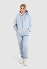 Ljusblå zip-up hoodie med framficka och dragsnören, kombinerad med matchande mjukisbyxor. Tyget verkar mjukt och texturerat.