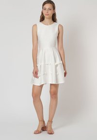 Vestido blanco sin mangas con un corsé ajustado, falda acampanada y tejido texturizado. Decorado con pequeños lazos de tela a lo largo del dobladillo.