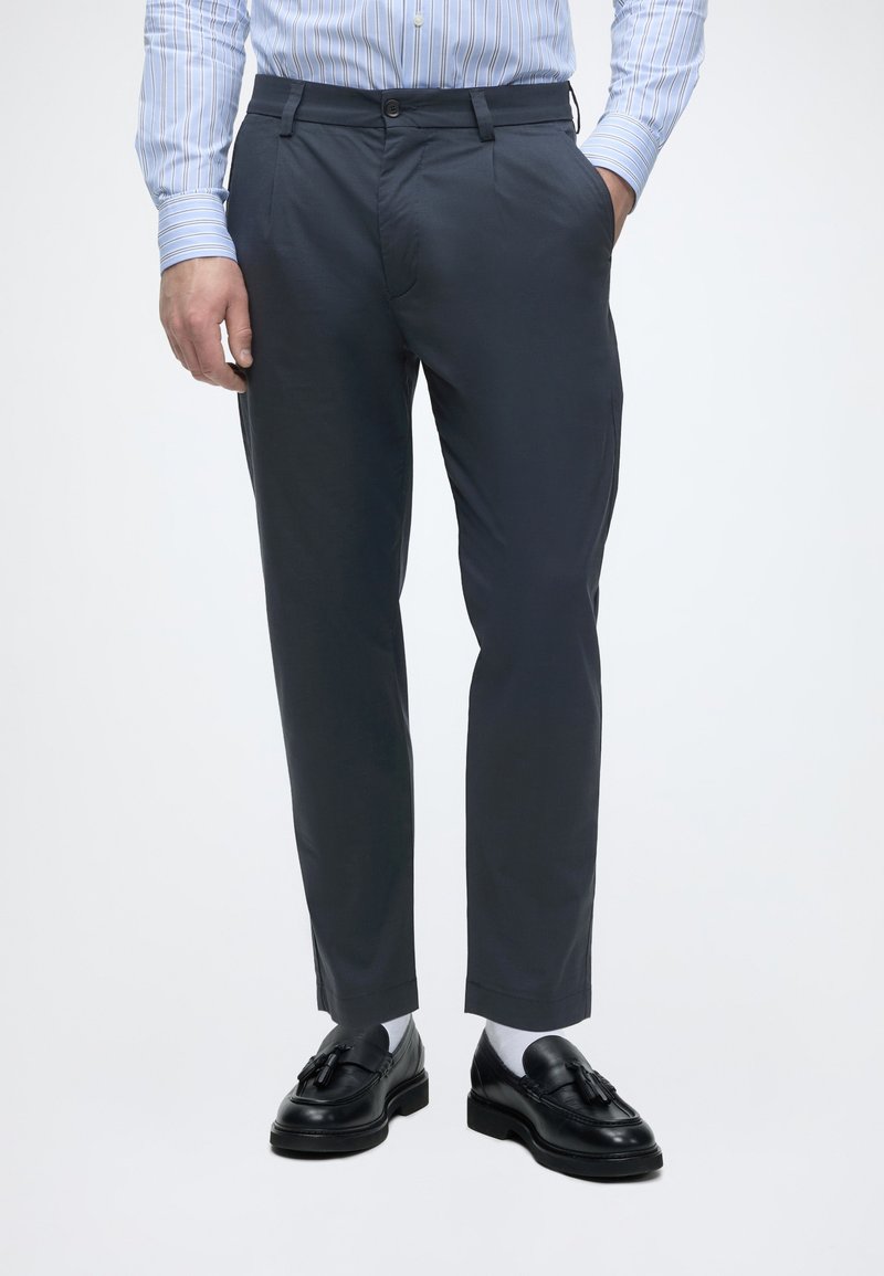 Homme portant un pantalon slim bleu marine, des chaussettes blanches, des mocassins noirs à pampilles et une chemise à rayures bleues, les mains dans les poches.