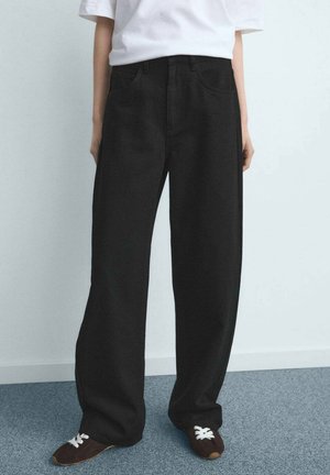 Personne portant un pantalon noir ample, un t-shirt blanc et des baskets marron à lacets blancs, debout sur un sol recouvert d'un tapis bleu, devant un mur blanc.