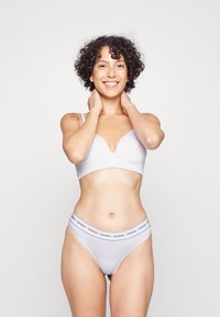 Soutien-gorge en tissu blanc avec un design en V et un string blanc assorti portant un logo "GUESS" noir sur la taille. Texture lisse et ajustée.