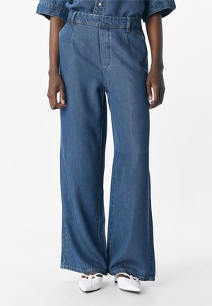 Person trägt hoch taillierte, weit geschnittene blaue Denim-Jeans mit einem Jeanshemd und weißen spitzen, niedrigen Absätzen.