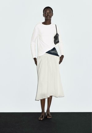 Witte lange mouwen shirt, donkerblauwe gebloemde taille-sjaal, vloeiende witte rok en open teen zwarte sandalen, gestyled met een kleine zwarte tas.