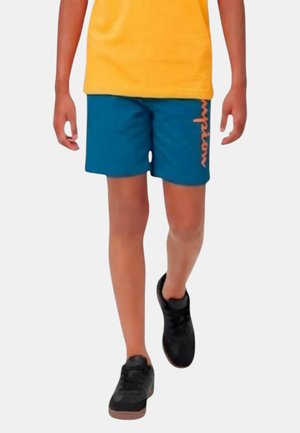 Bambino che indossa una maglietta gialla, pantaloncini blu con scritta arancione e scarpe da ginnastica nere che cammina su uno sfondo chiaro uniforme.