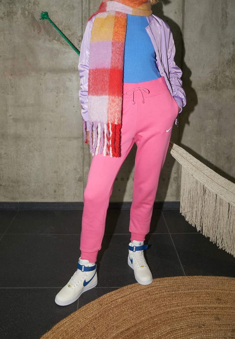 Roze sweatpants met een trekkoord, paarse bomberjack, blauwe geribbelde top en een gekleurde geruit sjaal; witte high-top sneakers met blauwe accenten.