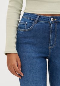 Vaqueros de mezclilla azules con cintura alta, diseño de cinco bolsillos, costuras visibles y una textura suave. Acompañados de una blusa de punto acanalado de color verde claro.