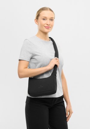 Mujer con cabello rubio, vestida con camiseta gris y pantalones negros, sosteniendo un bolso negro de hombro sobre un fondo blanco liso.