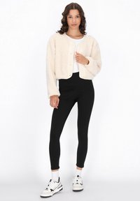 Giacca croppata di colore crema, morbida e pelosa, con scollo rotondo e chiusura a bottoni, abbinata a leggings neri e scarpe da ginnastica bianche. Texture liscia, stile casual.