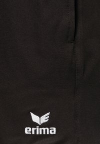 Pantalones cortos deportivos negros con una textura suave y un logo bordado en blanco. Incluyen un bolsillo lateral y un diseño sencillo sin patrones.