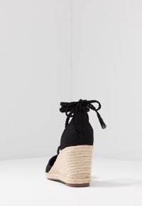 Černé espadrilové sandály na klínu s jute rope podpatkem, texturovaným textilem na svršku a pletenými řemínky okolo kotníků s střapci.