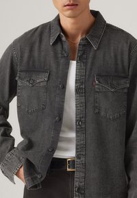 Camisa de mezclilla gris oscuro con dos bolsillos en el pecho, cierre de botones y una textura ligeramente rugosa. Llevada sobre una camiseta de tirantes blanca. Cinturón negro visible.
