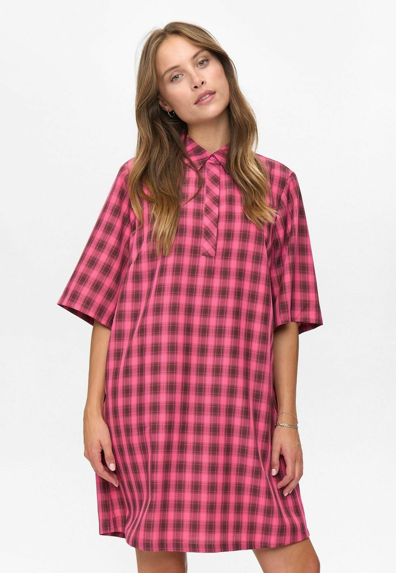 Jonge vrouw met lang haar, gekleed in een losvallende roze en donkere geruite jurk met korte mouwen, die tegen een effen witte achtergrond staat.